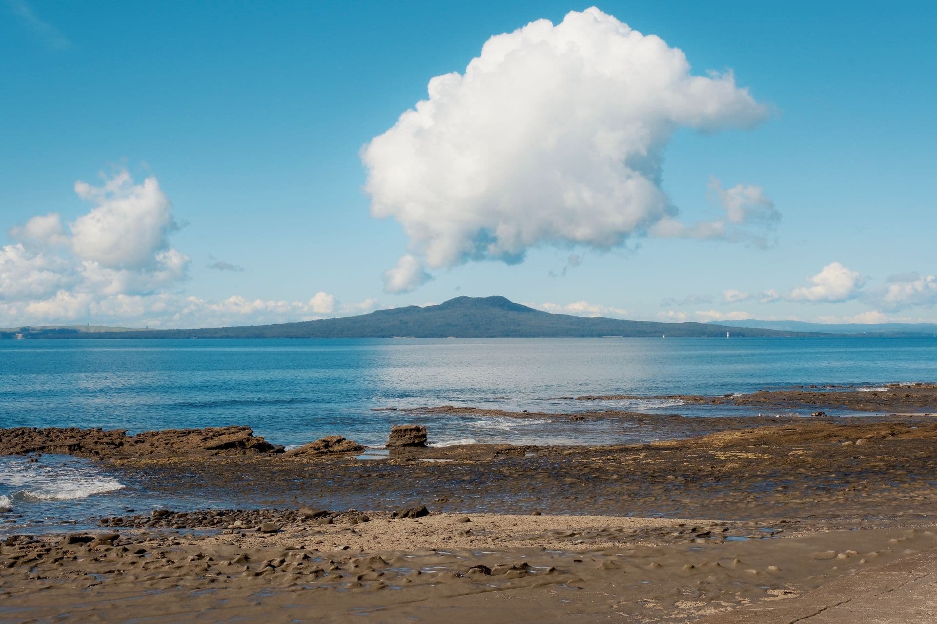 Rangitoto Island across calm blue water from a rocky Auckland beach under a large white cloud, New Zealand