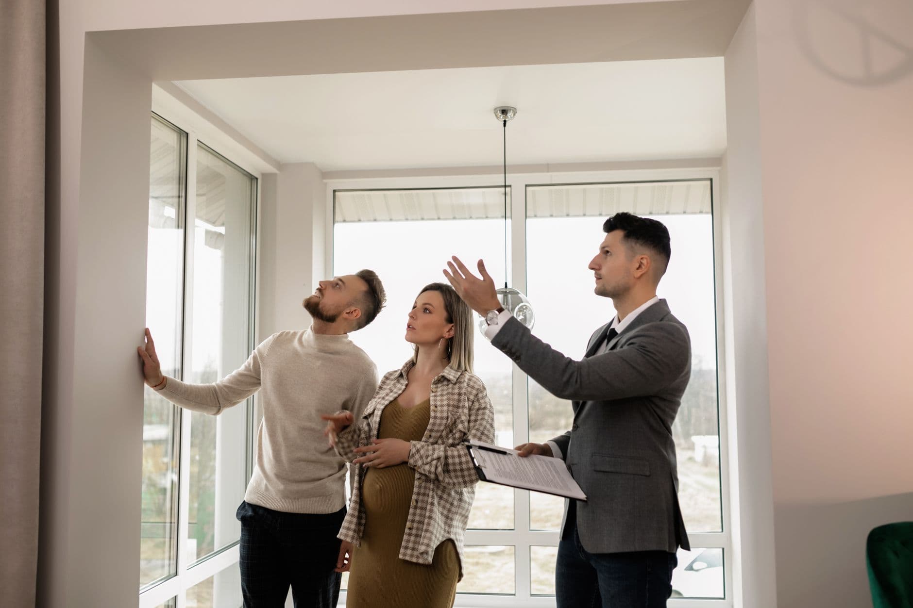 A couple looking up at a light fixture while viewing a bright, window-filled room with a real estate agent