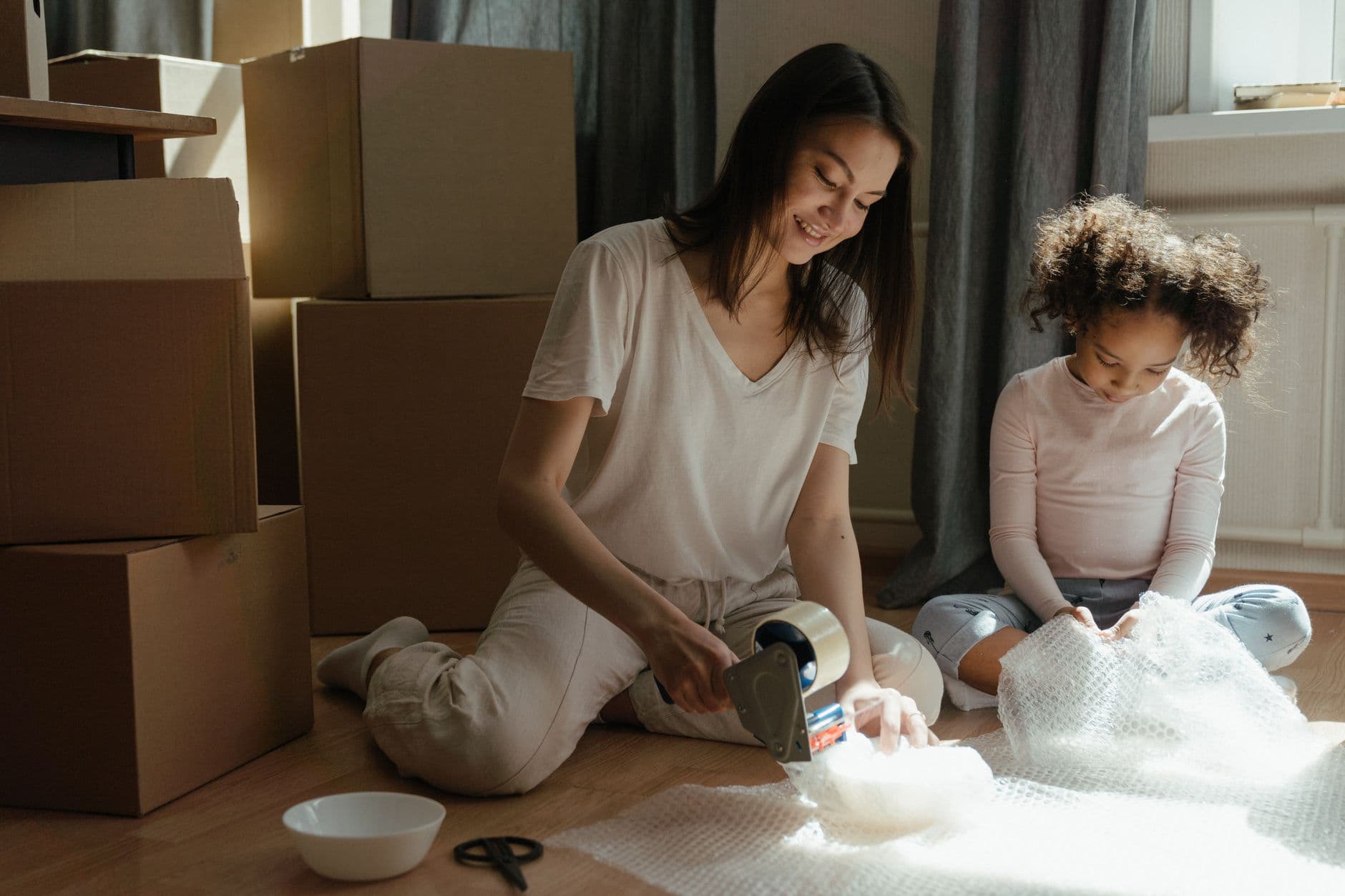 A mother and daughter sitting on the floor together, wrapping belongings in packing paper in a sunlit room